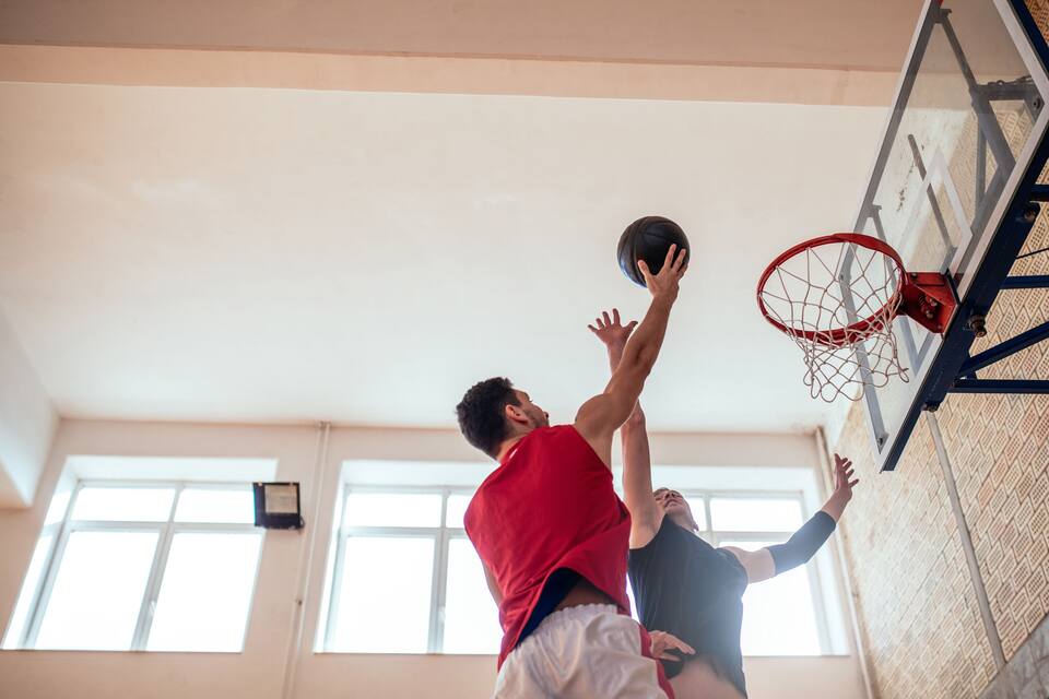 To personer spiller basketball i en indendørs hal med en basketballkurv og vinduer i baggrunden.