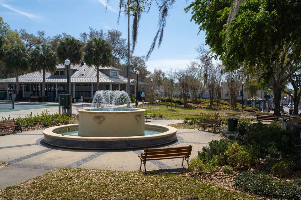 Um parque com uma fonte circular, bancos e um edifício ao fundo.