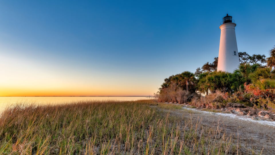 A white lighthouse with a black top, surrounded by grass and trees, at sunset.
