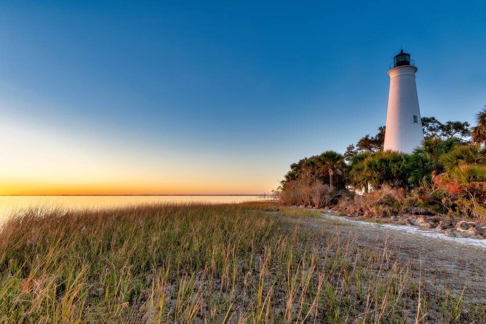 Um farol branco com topo preto, rodeado por grama e árvores, ao pôr do sol.