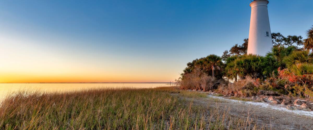 A white lighthouse with a black top, surrounded by grass and trees, at sunset.
