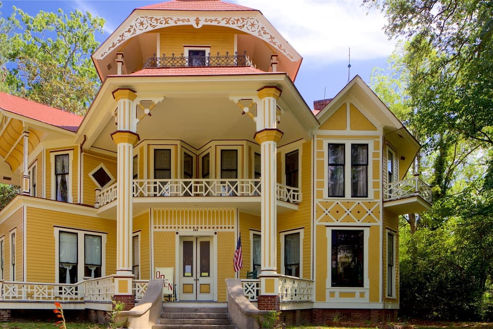 Una casa de estilo victoriano amarilla, con techo rojo, detalles blancos y un balcón.