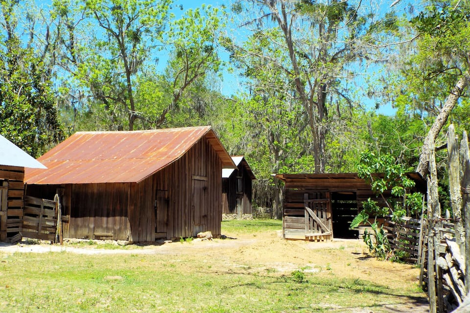 A rustic wooden barn with a rusted roof, surrounded by trees and a wooden fence.