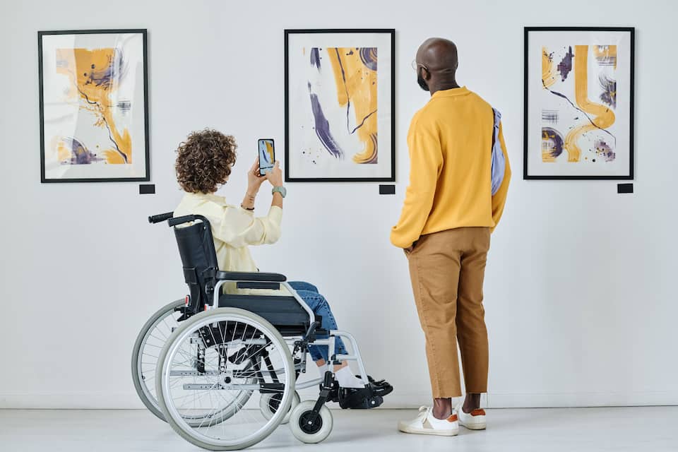 A person in a wheelchair and another person standing, both looking at framed abstract art on a white wall.