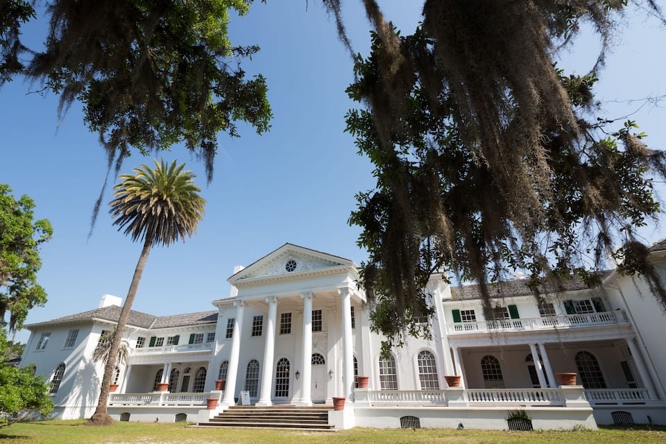 Un gran edificio de estilo colonial blanco con un pórtico central y columnas, rodeado de exuberante vegetación y una palmera.