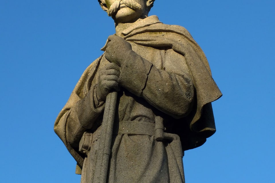 Estatua de piedra de una figura histórica con una túnica larga y un sombrero, sobre un cielo azul despejado.