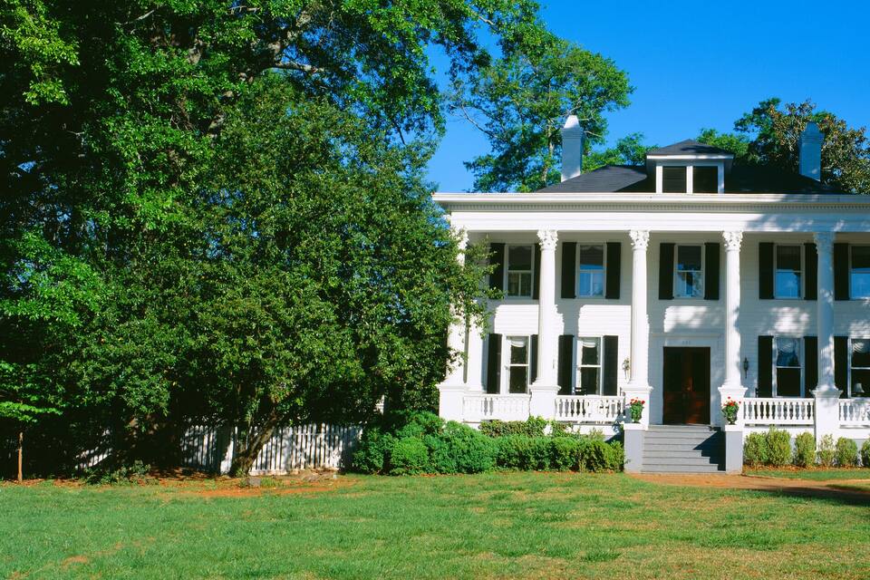 A white colonial-style house with a front porch, surrounded by lush green trees.
