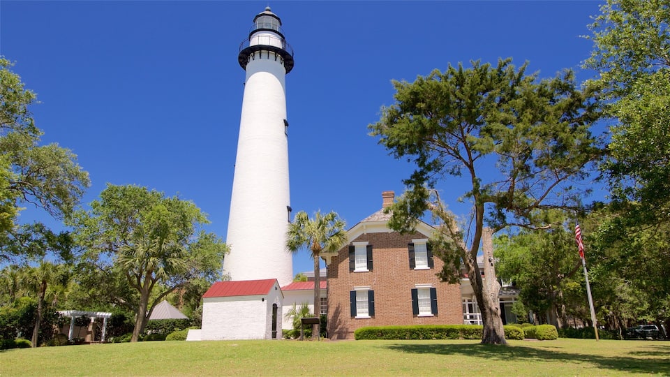 A tall white lighthouse with a red roof, surrounded by trees and a brick building.
