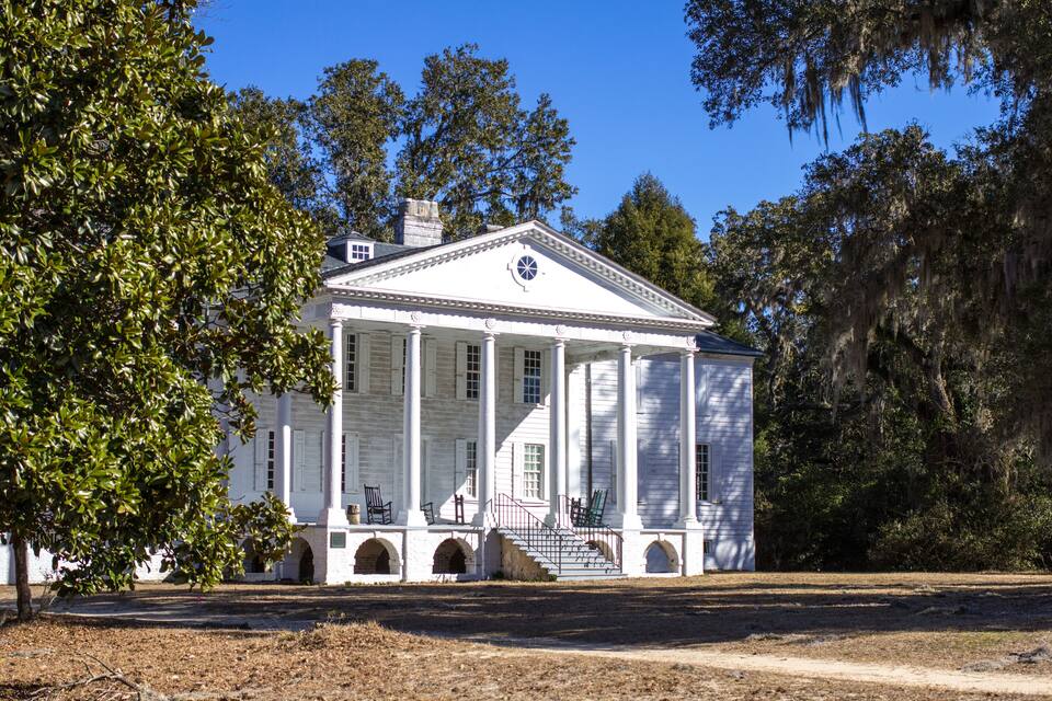 A white colonial-style building with columns and a porch, surrounded by trees and a clear sky.