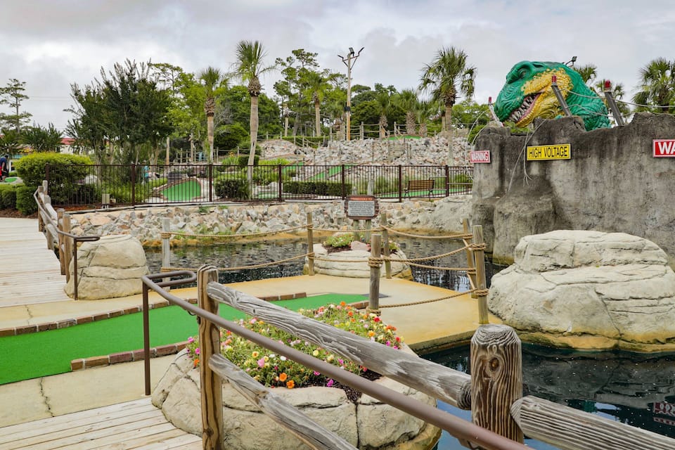 Atracció d'un parc temàtic aquàtic amb una estàtua de dinosaure verd, senyals d'advertència i un pont de fusta sobre un estany.
