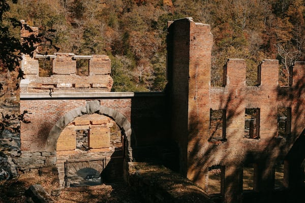 An abandoned brick structure with arched openings and a tall chimney amidst a forested hillside.