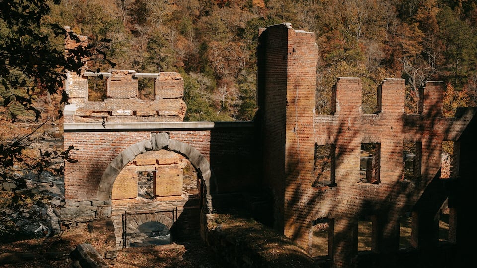 An abandoned brick structure with arched openings and a tall chimney amidst a forested hillside.