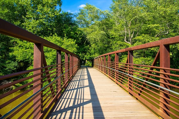 a long metal rust colored bridge over the silky brown waters of Little River surrounded by lush green trees, grass and plants with blue sky and clouds at Olde Rope Mill Park in Woodstock Georgia