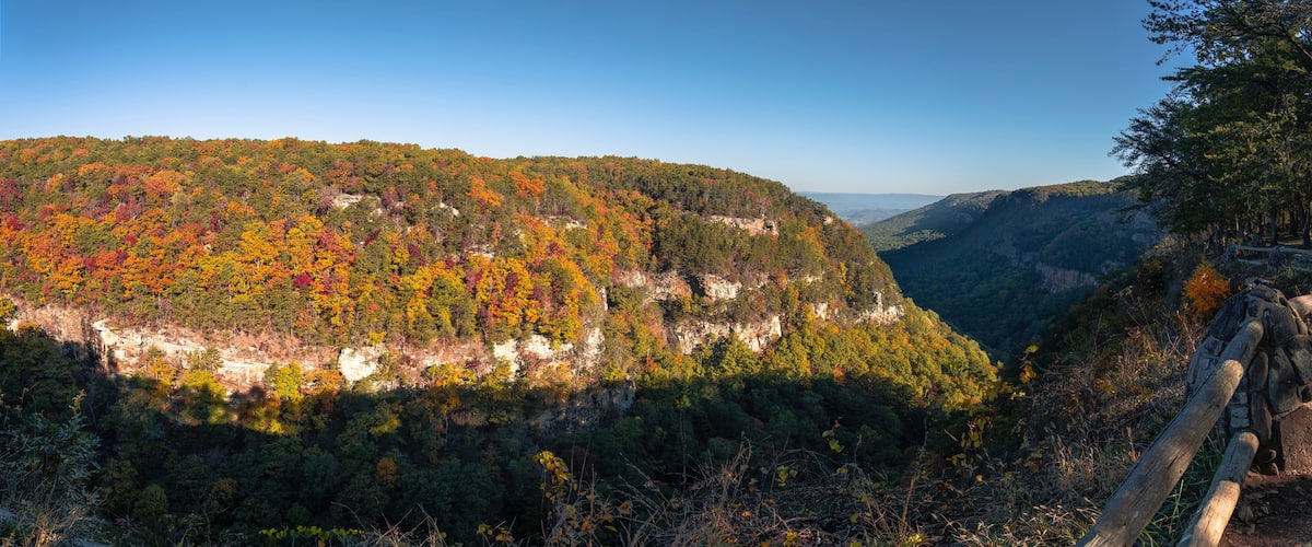 A forested mountain with autumn foliage, a rocky cliff, and a wooden fence.