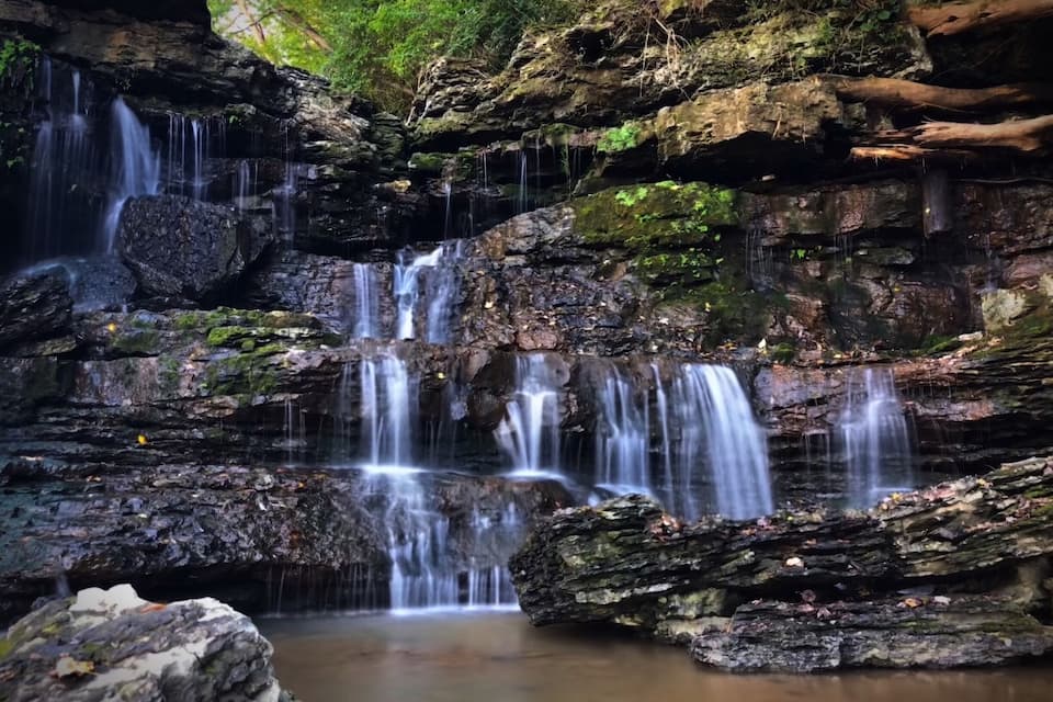 A multi-tiered waterfall cascading over rocky terrain with moss-covered stones and a calm pool at the base.