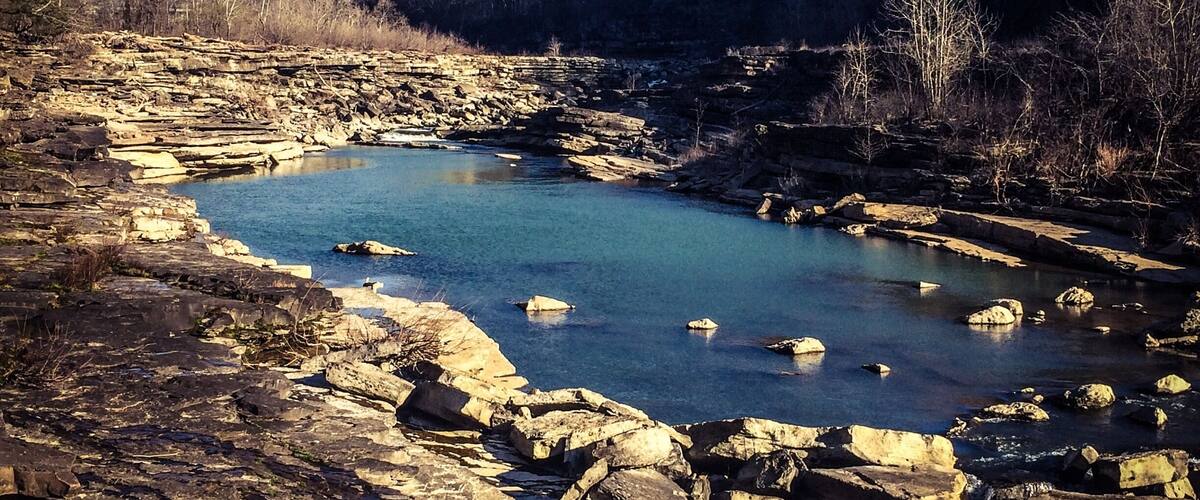 A rocky riverbed with clear water, surrounded by forested hills.