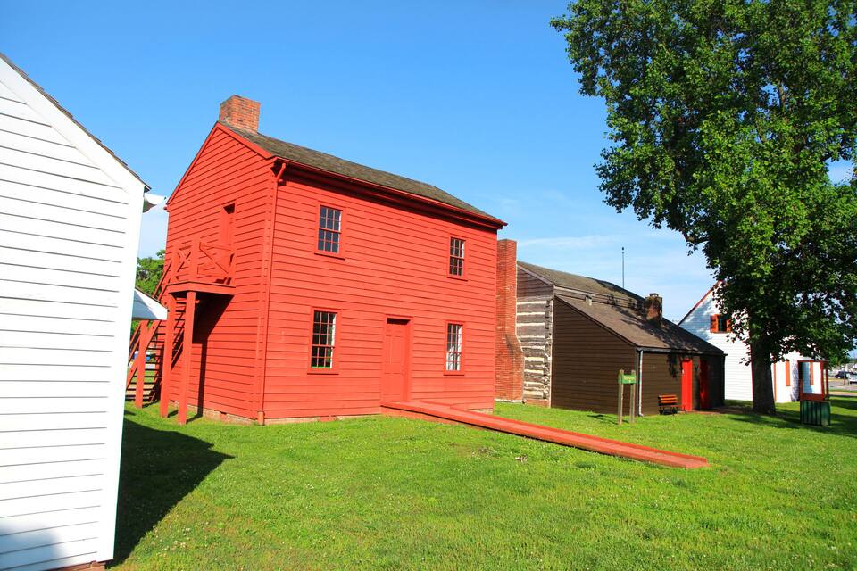 A red wooden house with a white adjacent building, a grassy yard, and a tree.
