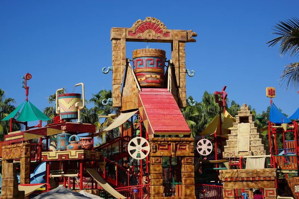 A colorful playground with a large red slide, a water fountain, and a decorative archway.