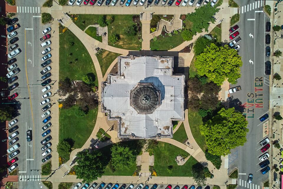 An aerial view of a city square with a central monument, surrounding greenery, and parked cars.