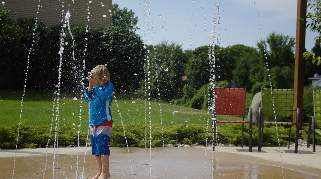 Little School Aged Caucasian Blond Boy in Red White and Blue Bathing Swim Suit Playing Outside Covering Face at Water Splash Pad with Beach Patio Chairs and Green Leaf Background