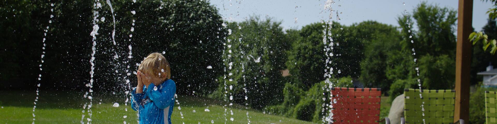 Little School Aged Caucasian Blond Boy in Red White and Blue Bathing Swim Suit Playing Outside Covering Face at Water Splash Pad with Beach Patio Chairs and Green Leaf Background