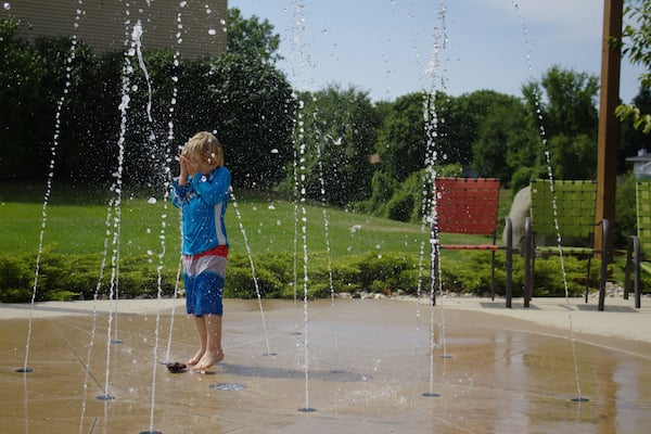 Little School Aged Caucasian Blond Boy in Red White and Blue Bathing Swim Suit Playing Outside Covering Face at Water Splash Pad with Beach Patio Chairs and Green Leaf Background