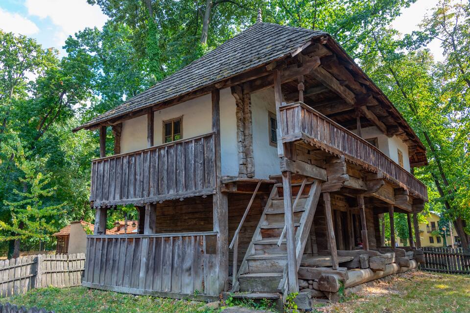 A rustic wooden house with a balcony, surrounded by lush greenery.