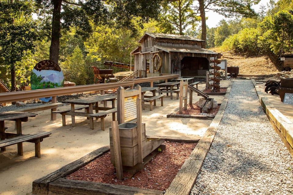 Un espace extérieur rustique comprenant des bancs en bois, des tables de pique-nique, une structure en bois portant l’inscription « Rocker Box » et un chemin de gravier.