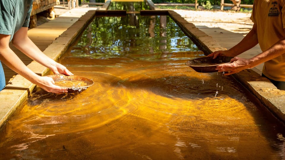 Two people holding bowls over a shallow, reflective water body with a wooden structure in the background.