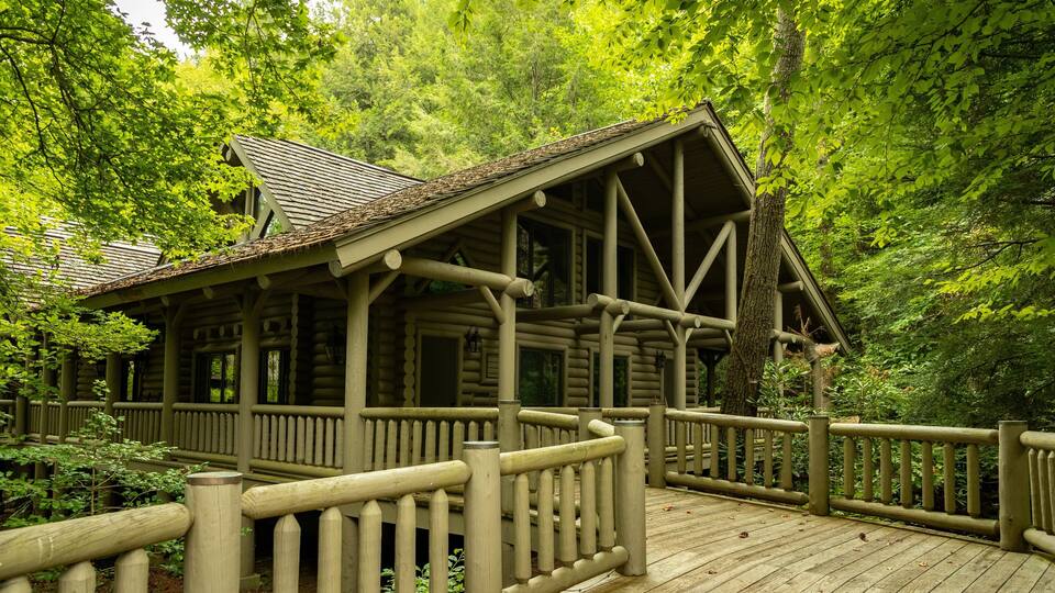 A wooden cabin with a porch and a wooden deck surrounded by dense forest.