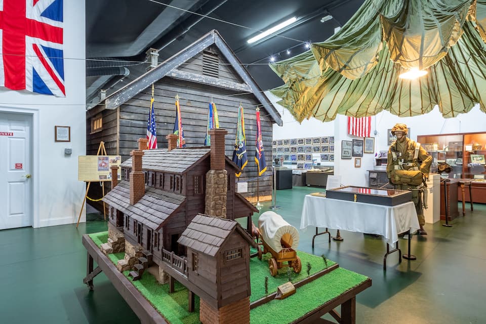 Una exposición histórica con una maqueta de casa, banderas y un maniquí de soldado.