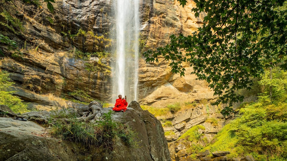 A waterfall cascading down a rocky cliff with a person meditating on a rock in the foreground.