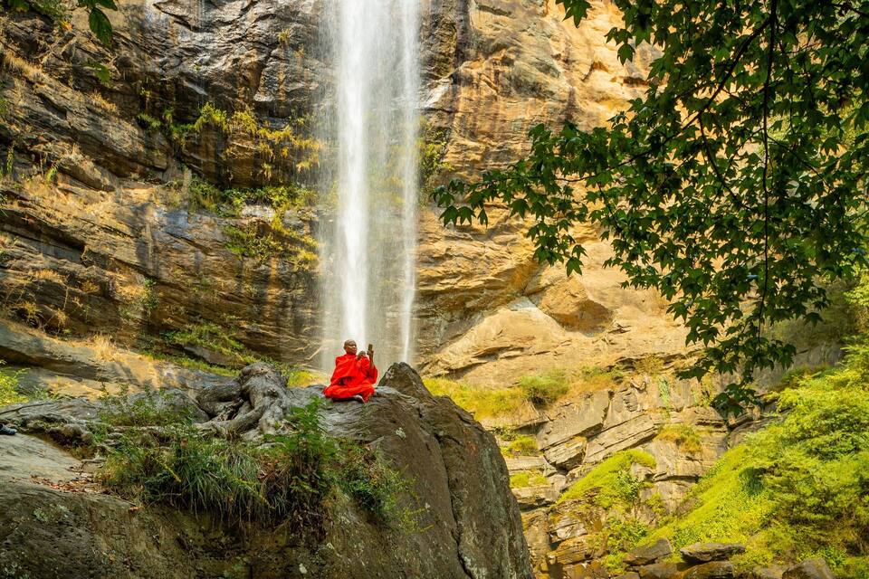 Una cascada se precipita por un acantilado rocoso, con una persona meditando sobre una roca al frente.