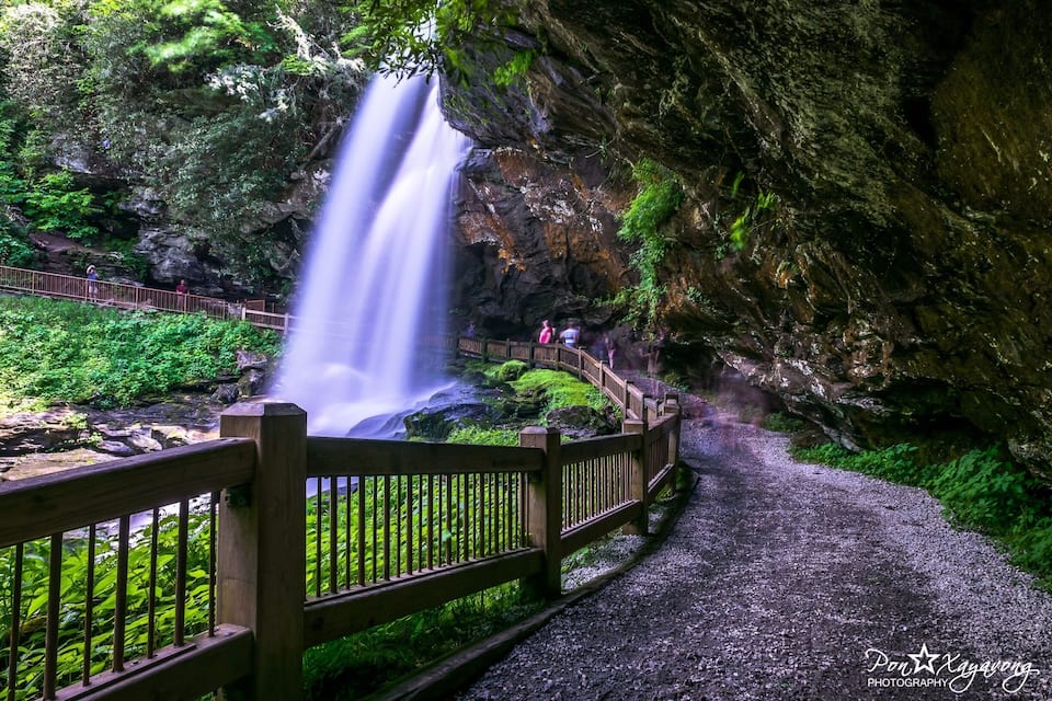 A waterfall cascading down a rocky cliff surrounded by lush greenery. A wooden walkway with a railing runs alongside the water.