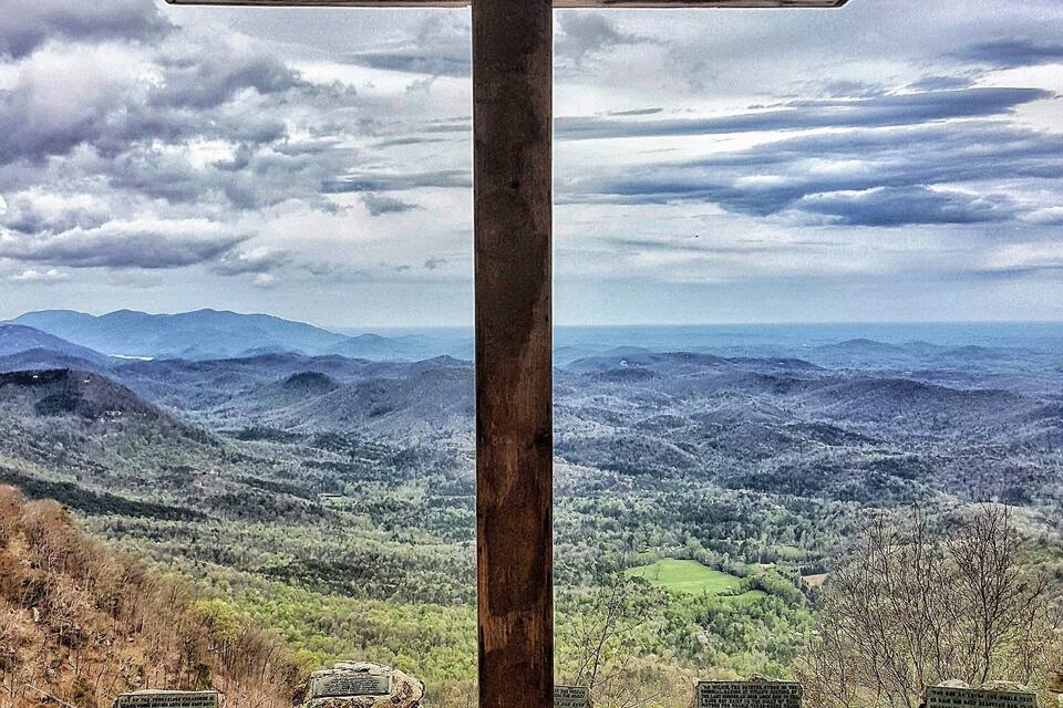 Una cruz de madera con inscripciones, frente a un paisaje montañoso bajo un cielo nublado.