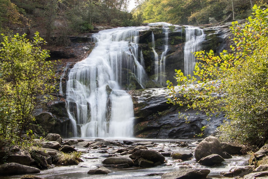Una cascada que se precipita sobre rocas en un área boscosa con abundante vegetación y un arroyo claro en la base.