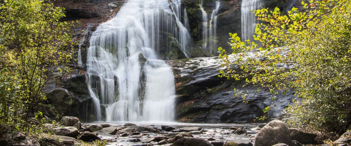 A waterfall cascading over rocks in a forested area with visible greenery and a clear stream at the base.