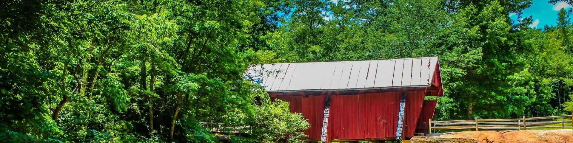 A red covered bridge over a stream, surrounded by lush greenery and trees.