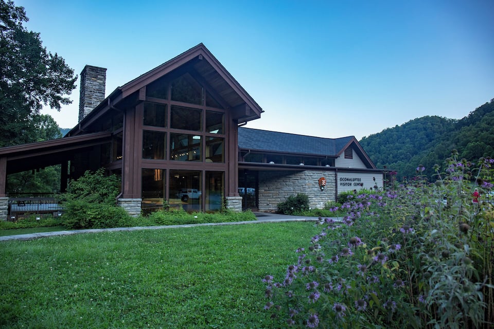 Un bâtiment moderne doté de grandes fenêtres et d’une façade en pierre, entouré d’une végétation luxuriante et d’un paysage vallonné.