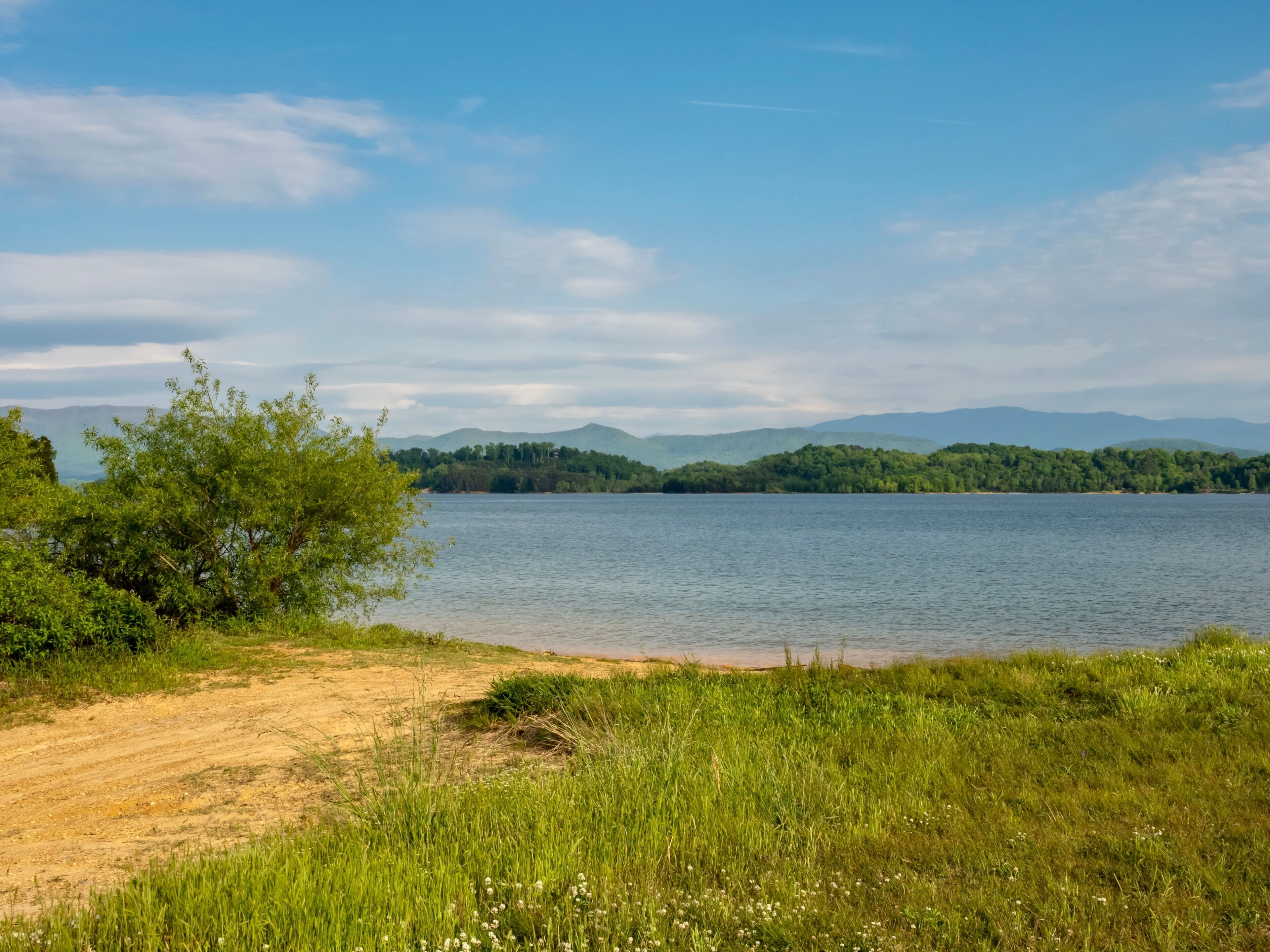 Douglas Lake in the spring in Tennessee with the Great Smoky Mountains in the background, the water and sandy shore with a blue sky filled with clouds in the distance.