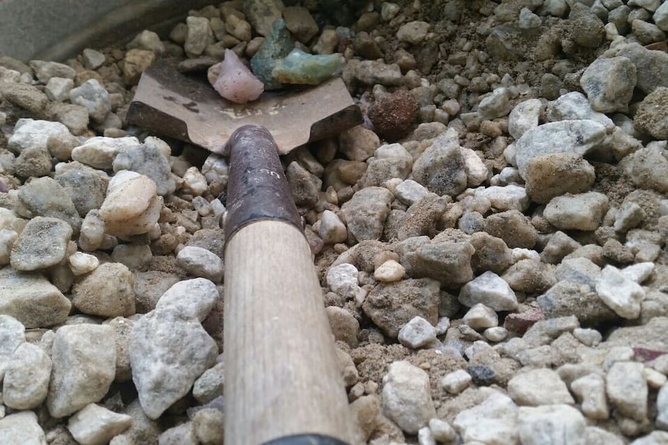 A garden tool with a wooden handle and a metal head resting on a bed of small rocks.