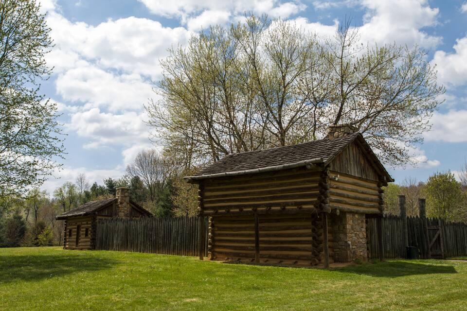 A rustic log cabin with a stone chimney, surrounded by a wooden fence and lush green grass.