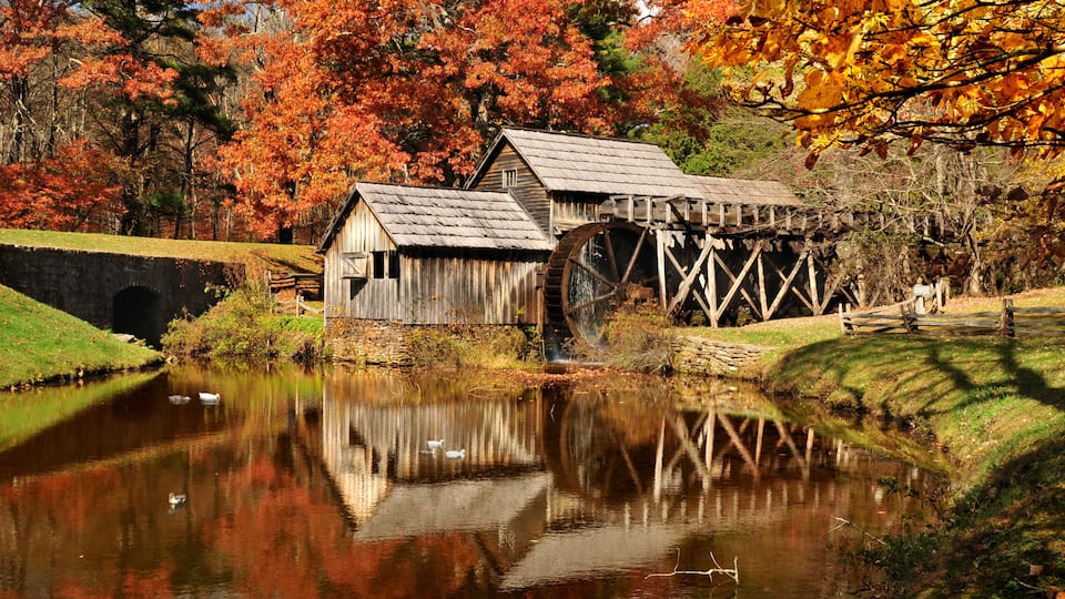 A rustic water mill with a wooden structure and a large water wheel, surrounded by a calm pond and autumn-colored trees.