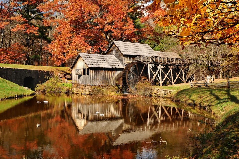 Eine rustikale Wassermühle mit Holzbau und einem großen Wasserrad, umgeben von einem stillen Teich und Bäumen in Herbstfarben.