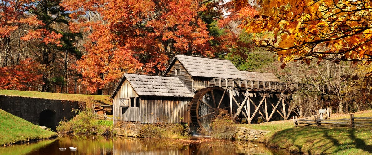 A rustic water mill with a wooden structure and a large water wheel, surrounded by a calm pond and autumn-colored trees.