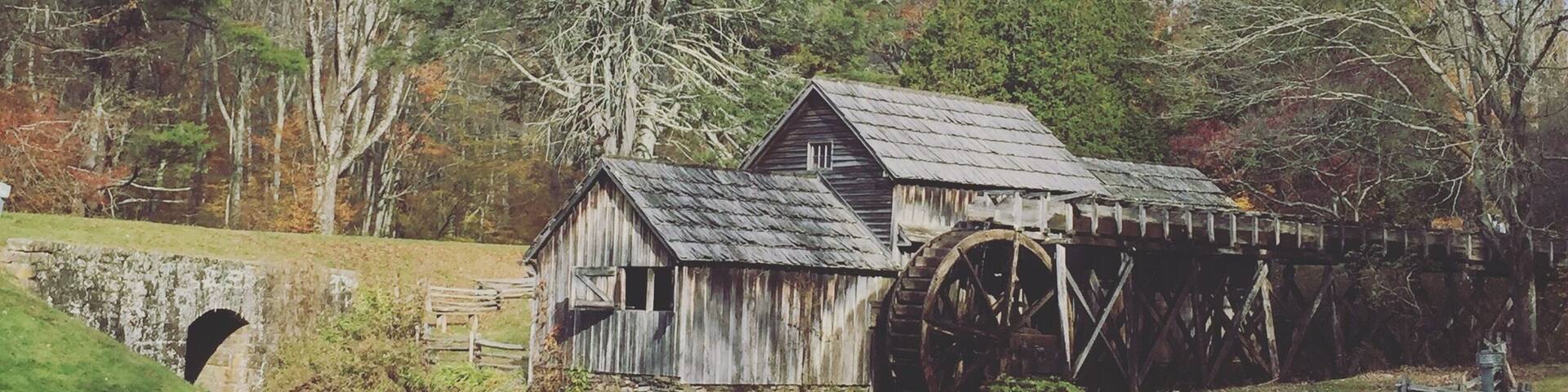A rustic water mill with a wooden structure and a large water wheel, surrounded by trees and a calm river.