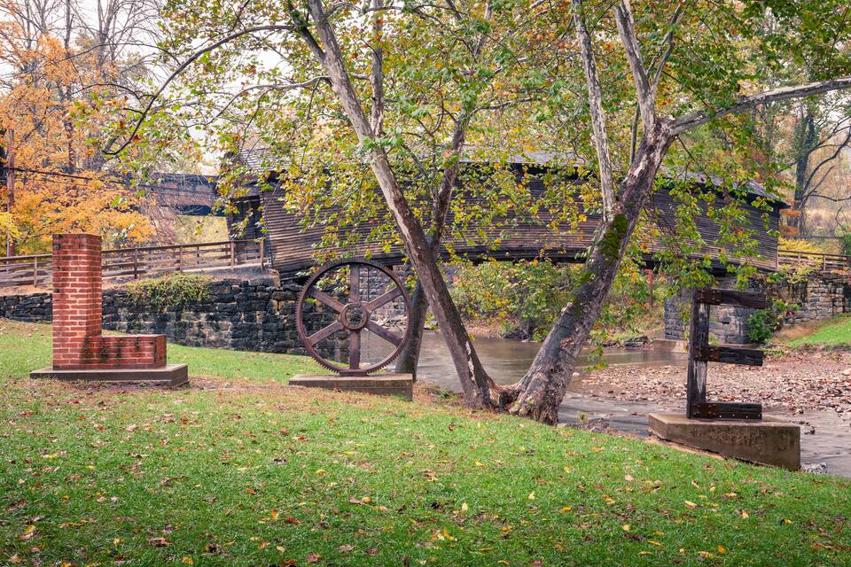A park with a brick structure, a large metal wheel, and a stone bridge over a river.
