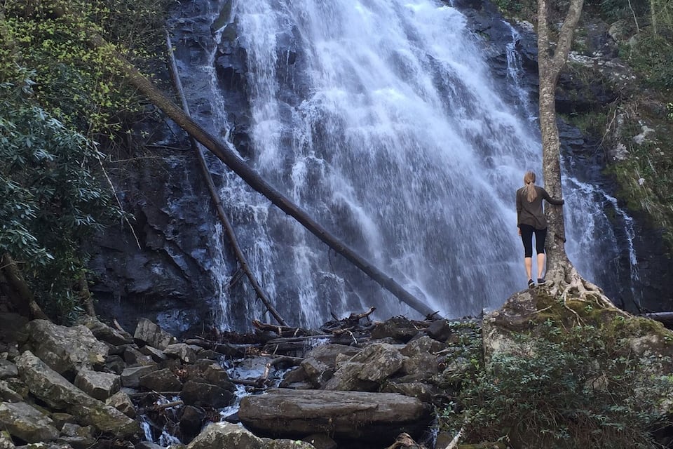 Una cascada en un área boscosa con una persona de pie sobre una repisa rocosa.