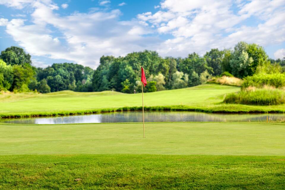Ein Golfplatz mit Teich, grünen Hügeln und einer roten Fahne.