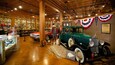 A vintage car exhibit with a green car and a red car on display, surrounded by bookshelves and American flags.